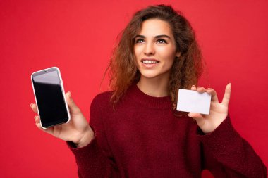 Samimi duyguları olan genç, kıvırcık esmer bir kadın arka plan duvarında yalnız başına kalmış, boş bir yerde, koyu kırmızı süveter giymiş, elinde kredi kartı cep telefonu olan, boş ekranlı bir cep telefonu tutan.