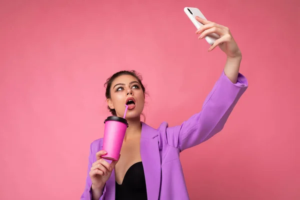 Close-up portrait of a beautiful young brunet woman in fashionable purple suit on a pink background in studio in a holding a popular phone and taking selfie photo and drinking through a straw beverage — Stock Photo, Image