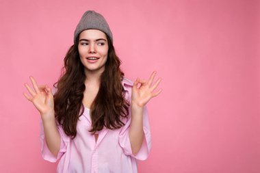 Güzel, mutlu, pozitif, kıvırcık, esmer, pembe tişörtlü, pembe arka planda izole edilmiş gri şapkalı bir kadın. Yoga konsepti