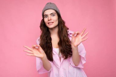 Pembe gömlekli, gri şapkalı, güzel, kıvırcık esmer kadının portresi. Pembe arka planda izole edilmiş ve güzel bir jest sergiliyor.