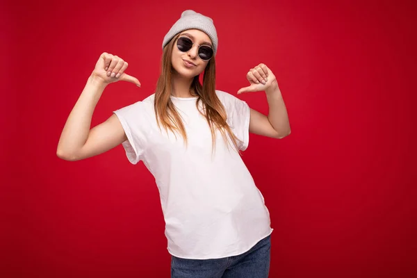 Foto de una joven rubia oscura positiva con emociones sinceras usando una camiseta blanca casual con espacio vacío para sombrero gris y gafas de sol aisladas sobre fondo rojo con espacio para copiar —  Fotos de Stock