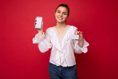 Beyaz bluz giyen, güzel Avrupalı, şık esmer bir kadın ve kırmızı arka planda izole edilmiş, samimi duygularla izole edilmiş şık bir şapka. Basit ve doğal bir şekilde kameraya bakıyor. Tutuluyor