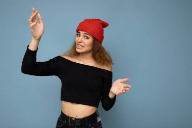 Genç, duygusal, tatminsiz, hoş, esmer, dalgalı saçlı, samimi duyguları olan, siyah bluz giyen, mavi arka planda izole edilmiş kırmızı şapkalı bir kadın.