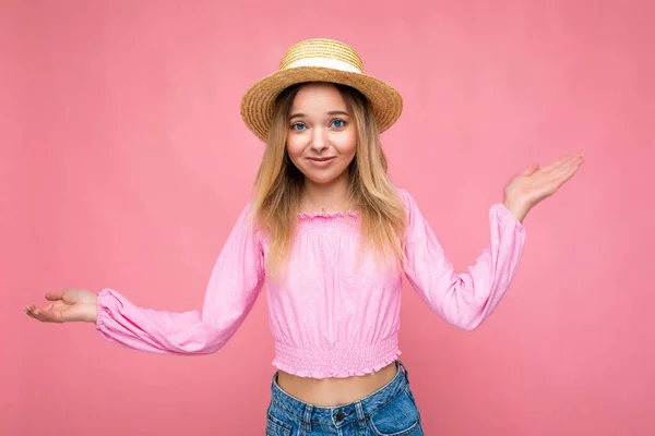 Фото фото молодой красивой милой счастливой блондинки, носящей стильный розовый топ и соломенную шляпу на розовом фоне с копировальным пространством. Концепция сомнений — стоковое фото