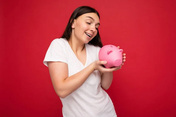 Rozkošné pozitivní úsměv mladá krásná okouzlující brunetka žena s upřímnými emocemi na sobě ležérní bílé tričko izolované přes červené pozadí s kopírovacím prostorem a držení růžové prasátko box. Moneybox — Stock fotografie