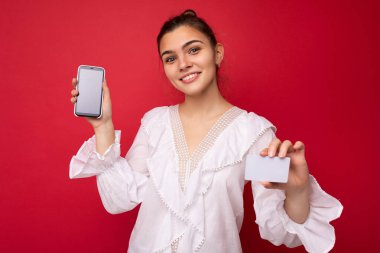 Beyaz bluz giyen, güzel Avrupalı, şık esmer bir kadın ve kırmızı arka planda izole edilmiş, samimi duygularla izole edilmiş şık bir şapka. Basit ve doğal bir şekilde kameraya bakıyor. Tutuluyor