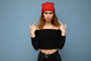 Genç, pozitif, esmer, kıvırcık saçlı, samimi duyguları olan, siyah bluzlu ve kırmızı şapkalı bir kadının fotoğrafı. Fotokopi alanı olan mavi arka planda izole edilmiş ve eğleniyor.