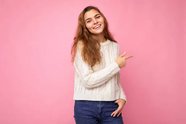 Молодая красивая европейская стильная брюнетка в белом зимнем свитере изолирована на розовом фоне с положительными искренними эмоциями. Простой и естественный взгляд на камеру. Указание пальцем на — стоковое фото