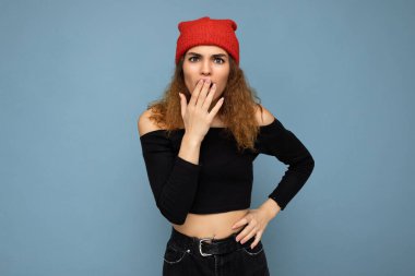 Genç duygusal şaşkınlık, hoş, rüzgarlı, esmer, samimi duyguları olan, siyah bluz giyen ve mavi arka planda izole edilmiş kırmızı şapkalı bir kadın.