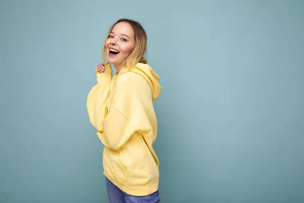 Joven mujer hermosa. Mujer de moda en sudadera hipster amarillo de moda. La hembra positiva muestra emociones faciales. Modelo divertido aislado sobre fondo azul con espacio libre —  Fotos de Stock