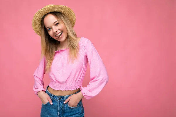 Retrato de una joven atractiva y sonriente mujer rubia hipster con elegante top rosa y sombrero de paja. Sexy persona femenina despreocupada posando aislado cerca de la pared rosa en el estudio. Modelo positivo con natural — Foto de Stock