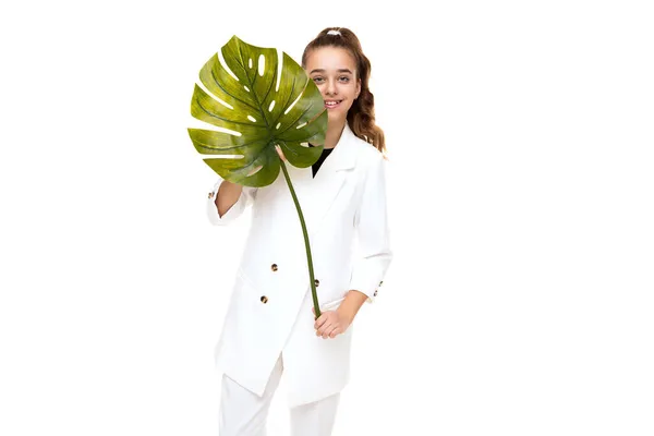 Foto van een mooie schattige schattige lachende brunette tienermeisje met een paardenstaart in een stijlvolle witte jas en witte broek met een groen groot blad van een tropische plant geïsoleerd op een witte achtergrond — Stockfoto