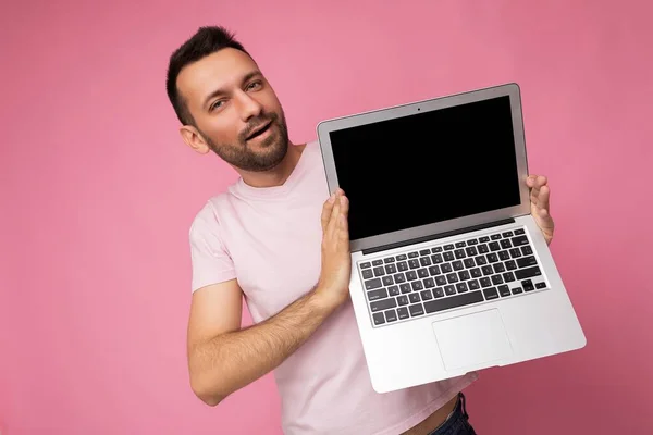 Hombre guapo moreno sosteniendo computadora portátil con monitor negro en blanco para maqueta mirando a la cámara en camiseta rosa aislada sobre fondo rosa con espacio libre para el texto —  Fotos de Stock