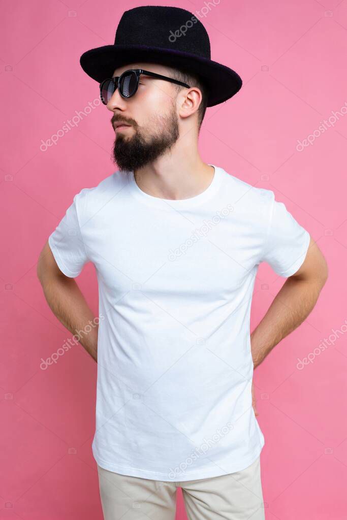 Vertical photo shot of confident brutal good looking young brunet bearded man wearing casual white t-shirt for mockup , sunglasses and stylish black hat poising isolated on pink background with empty