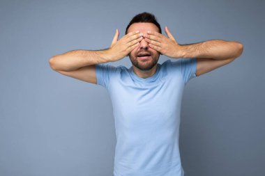 Yetişkin, yakışıklı, sakalsız, samimi duyguları olan, mavi tişörtlü, sakalsız bir adamın fotoğrafı. Mavi arka plan duvarına izole edilmiş, boş bir alan ve elleriyle gözlerini kapatan bir adam.