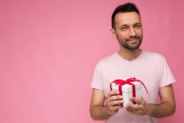 Yakışıklı, kendine güvenen esmer bir adam elinde kırmızı kurdeleli beyaz bir hediye kutusu tutuyor. Pembe tişörtlü, pembe arka planda izole edilmiş ve mesaj için boş yeri var.