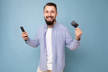 Güzel görünüşlü, çekici, gülümseyen, tıraşsız, mavi gömlekli, beyaz tişörtlü genç bir adamın fotoğrafı. Mavi arka plan duvarında, kredi kartı ve cep telefonu ile izole edilmiş.