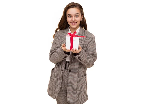Φωτογραφία από όμορφο χαριτωμένο αρκετά χαρούμενος χαμογελαστή μελαχρινή έφηβη κοπέλα με μακριά μαλλιά σε κομψό καρό σακάκι, καρό παντελόνι και λευκό πουκάμισο κρατά λευκό κουτί με κόκκινη κορδέλα με ένα δώρο που απομονώνονται σε λευκό — Φωτογραφία Αρχείου