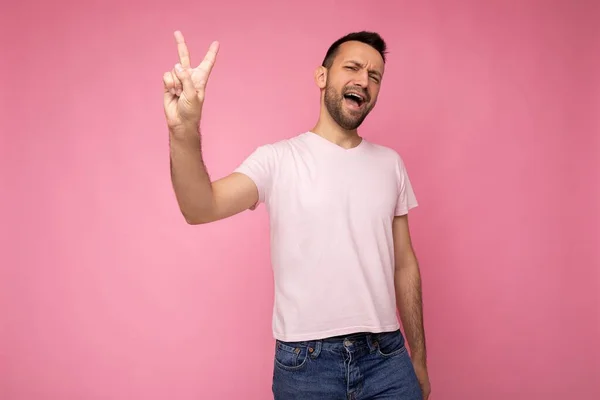 핑크 색 티셔츠를 입은 잘생긴 감정적 긍정적 인 갈색 남성의 사진은 텍스트를 복사할 공간 과 두 손가락으로 평화 로운 몸짓을 하는 핑크 배경에 고립되어 있다 — 스톡 사진