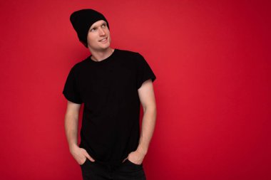 Yakışıklı, pozitif, düşünceli yetişkin bir adamın kırmızı arka plan duvarının üzerinde dikilip siyah t-shirt giyip siyah şapka giyip yan tarafa bakıp şöyle düşünmesi.