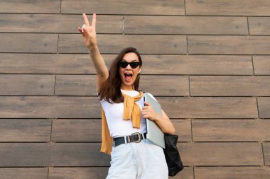 Güzel, gülümseyen, genç bir esmer kadın elinde bilgisayar dizüstü bilgisayar ve güneş gözlüğü olan kameraya bakıyor. Barış jesti ve dili beyaz tişört ve açık mavi kot pantolonla sokakta görülüyor.