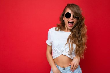 Güzel, pozitif sarışın bir kadının, günlük kıyafetler giydiği ve renkli arka planda izole edilmiş güneş gözlüğü taktığı fotoğraf. Kameraya bakıyor.