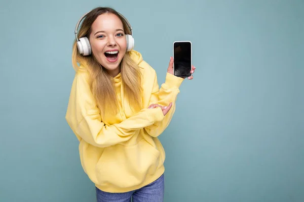 Atractiva mujer joven sonriente positiva con elegante atuendo casual aislado en la pared de fondo de colores que sostiene y muestra el teléfono móvil con pantalla vacía para recortar el uso de bluetooth blanco — Foto de Stock