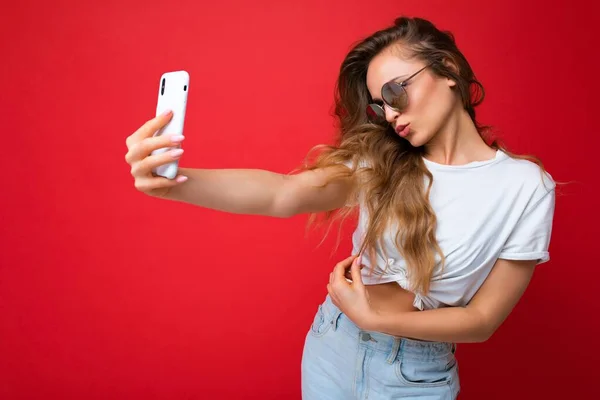 Foto de primer plano de la increíble hermosa mujer rubia joven sosteniendo el teléfono móvil tomando foto selfie utilizando la cámara del teléfono inteligente con gafas de sol traje elegante todos los días aislado sobre fondo de pared colorido — Foto de Stock