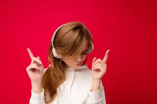 Atractiva mujer rubia emocional positiva con capucha blanca aislada sobre fondo colorido con auriculares bluetooth inalámbricos blancos escuchando música y bailando — Foto de Stock
