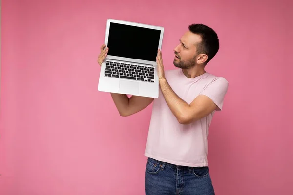 Hombre guapo sorprendido y sorprendido sosteniendo ordenador portátil mirando netbook pantalla en camiseta sobre fondo rosa aislado —  Fotos de Stock
