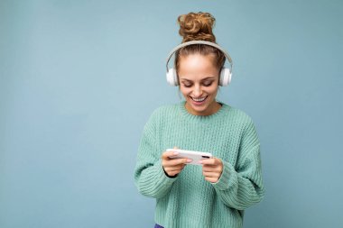 Mavi arka plan üzerine izole edilmiş mavi süveter giyen, beyaz bluetooth kulaklık takan, müzik dinleyen ve internet üzerinden cep telefonu kullanan güzel, pozitif bir genç kadın.