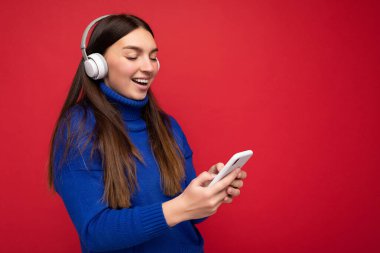 Güzel, neşeli, güler yüzlü, şık kıyafetler giyen, arka plan duvarından izole edilmiş, cep telefonu kullanan, beyaz bluetooth kulaklık takan ve müzik dinleyen genç bir kadının fotoğrafı.