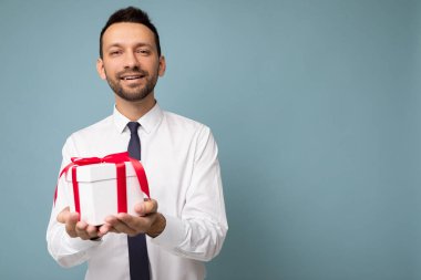 Yakışıklı, pozitif esmer, sakalsız, mavi arka plan duvarında izole edilmiş beyaz gömlekli, kırmızı kurdeleli ve kameraya bakan genç bir adamın fotoğrafı.