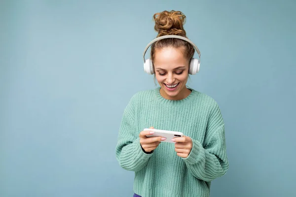 파란 스웨터를 입고 푸른 배경에 고립된 아름다운 양성의 젊은 여성 이 흰색 블루투스 무선 이어폰을 착용하고 음악을 듣고 온라인으로 전화 통화를 하고 있다. — 스톡 사진