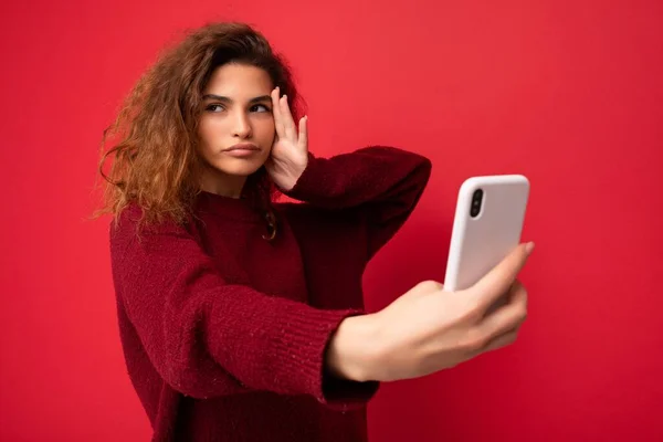 Bela jovem concentrada séria com cabelo encaracolado vestindo camisola vermelha escura isolada na parede de fundo vermelho segurando e usando telefone inteligente olhando para o lado e tirando selfie — Fotografia de Stock