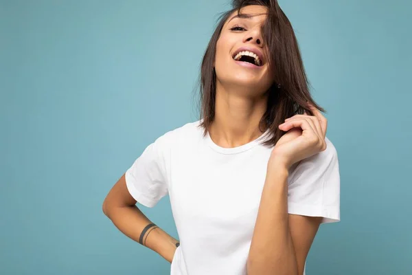 若い美しい幸せな笑顔のブルネットの女性の写真は、モックアップのための空のスペースと流行の白いTシャツを着ています。フリースペースとスタジオで青い壁の近くに隔離されたセックス屈託のない女性の人 — ストック写真