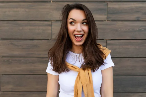 Portret van een succesvolle glimlachende vrolijke jonge brunet vrouw met casual wit t-shirt en jeans met gele trui in de buurt van bruine muur in de straat en veel plezier — Stockfoto