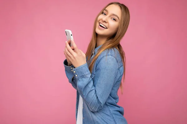 밝은 청색 청바지를 입은 아름다운 금발 여성 이 손에 핑크 색종이를 들고 고립되어 있고 인터넷에서 휴대 전화 서핑을 하며 카메라를 보고 있다. — 스톡 사진