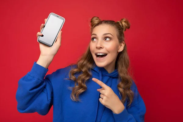 Jolie jeune femme blonde souriante portant un élégant sweat à capuche bleu isolé sur fond rouge avec espace de copie tenant smartphone montrant téléphone en main avec écran vide pointant vers gadjet — Photo