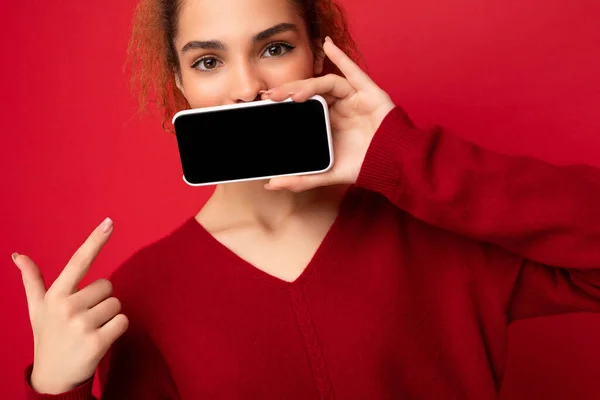 Closeup photo of young woman wearing dark red sweater isolated over red background holding smartphone and showing mobile phone screen with copy space for cutout looking at camera and pointing at — Stock Photo, Image