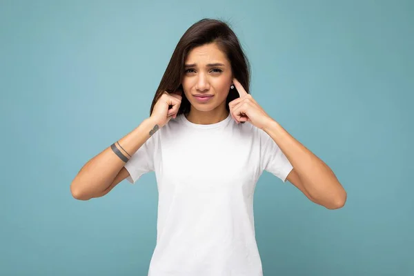 Portrait of young beautiful brunette woman with sincere emotions wearing casual white t-shirt for mockup isolated on blue background with empty space and covering ears with hands