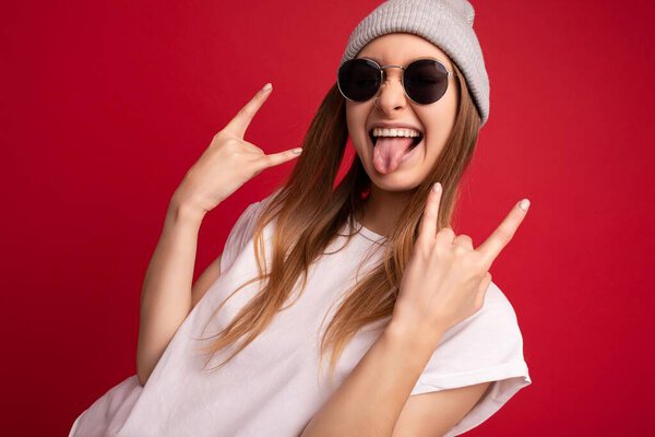 Portrait photo of young emotional positive happy funny laughing beautiful attractive dark blonde woman wearing casual white t-shirt with empty space for mockup, grey hat and sunglasses isolated over