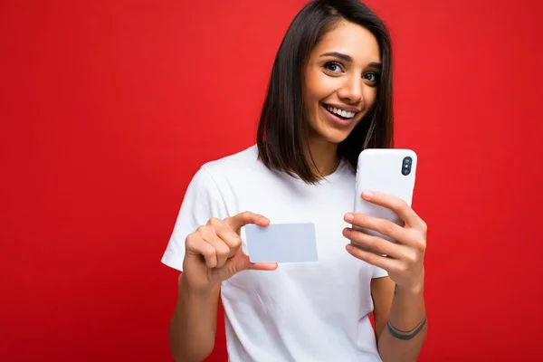 Prachtige vrolijke glimlachende jonge vrouw met kort donker brunet kapsel met witte plastic bankkaart voor mockup en mobiele telefoon voor online betaling met wit t-shirt geïsoleerd over rode muur — Stockfoto
