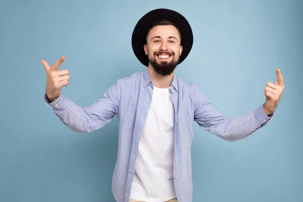 Photo shot of positive happy smiling good looking young brunet bearded man wearing casual blue shirt and white t-shirt and stylish black hat poising isolated on blue background with free space for — Stock Photo, Image