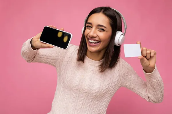 Gyönyörű vonzó pozitív boldog mosolygós fiatal nő visel elegáns alkalmi ruhát és fehér vezeték nélküli fejhallgató elszigetelt színes háttér fal gazdaság műanyag hitelkártya és mutatja a mobil — Stock Fotó