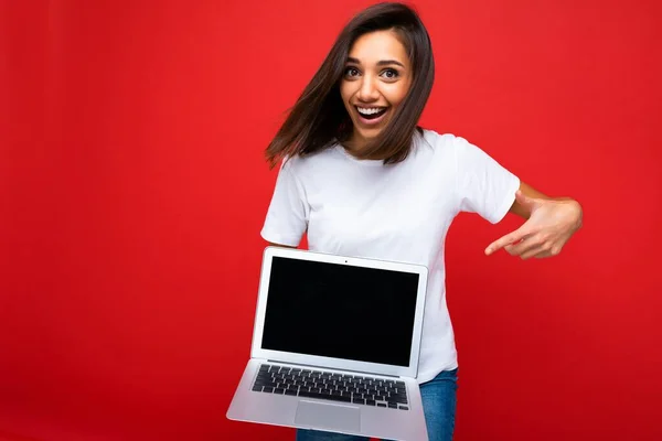 Όμορφη χαρούμενη νεαρή γυναίκα με κοντά σκούρα καστανά μαλλιά κρατώντας φορητό υπολογιστή με μαύρη λευκή οθόνη για mockup φορώντας λευκό t-shirt και τζιν που απομονώνονται πάνω από τον κόκκινο τοίχο — Φωτογραφία Αρχείου