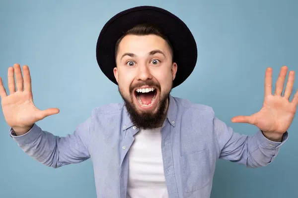 Närbild foto skott av positiva roliga glada glada glada leende snygg ung brunett skäggig man bär casual blå skjorta och vit t-shirt och elegant svart hatt balansera isolerad på blå bakgrund — Stockfoto