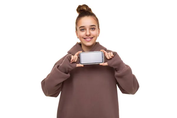 Foto de uma linda garota bonita com cabelo escuro e um pão com capuz marrom com um capuz segurando um telefone celular com uma tela preta em branco isolada em um fundo branco com espaço livre para texto — Fotografia de Stock