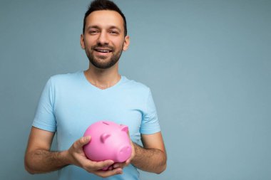 Mutlu, sevimli, gülümseyen, sakalsız, sakalsız, samimi duyguları olan, mavi tişörtlü, mavi arka plan duvarına izole edilmiş ve elinde pembe domuzcuk tutan yakışıklı bir adamın fotoğrafı.