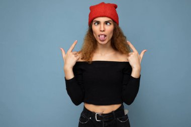 Genç, pozitif, esmer, kıvırcık saçlı, samimi duyguları olan, siyah bluzlu ve kırmızı şapkalı bir kadının fotoğrafı. Mavi arka planda izole edilmiş.